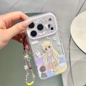 Peut inclure: Une coque de téléphone transparente avec un contour d'appareil photo rose clair, illustrée d'un personnage blond entouré de lapins blancs et d'un chat noir. Elle est accompagnée d'une dragonne perlée avec des perles colorées et des fleurs.