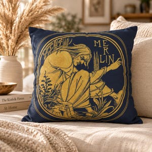 May include: A square throw pillow with a dark navy blue background and a gold-colored graphic print. The design features a seated figure within an oval frame, surrounded by foliage, with the text MERLIN printed in a vertical gold font.