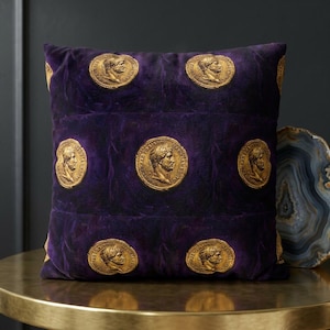 Op de afbeelding: Vierkantig decoratief kussen in een diepe paarse tint, versierd met een herhalend patroon van goudkleurige Romeinse muntontwerpen. Het kussen is gemaakt van een zacht, fluwelig materiaal en ligt op een goudkleurige tafel.
