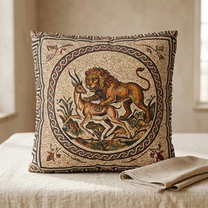 May include: Square decorative pillow with a mosaic design featuring a lion attacking a deer. The pillow has a beige background with brown and tan accents, and a decorative border. A folded beige napkin rests on the table.