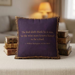 May include: A square purple throw pillow featuring a gold-colored ornate frame border and the printed text: The fool doth think he is wise, but the wise man knows himself to be a fool. — William Shakespeare, As You Like It.