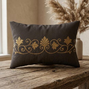 May include: A rectangular dark gray throw pillow featuring a horizontal band of metallic gold embroidery. The embroidery displays a repeating pattern of stylized acanthus leaves and scrolling vines centered across the front of the pillow.