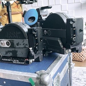 May include: A professional black Arriflex 16SRII film camera with a blue felt cover, resting on a blue case with silver metal corners. The camera has the text "ARR" and "ARRFLEX 16SRII" printed on it.