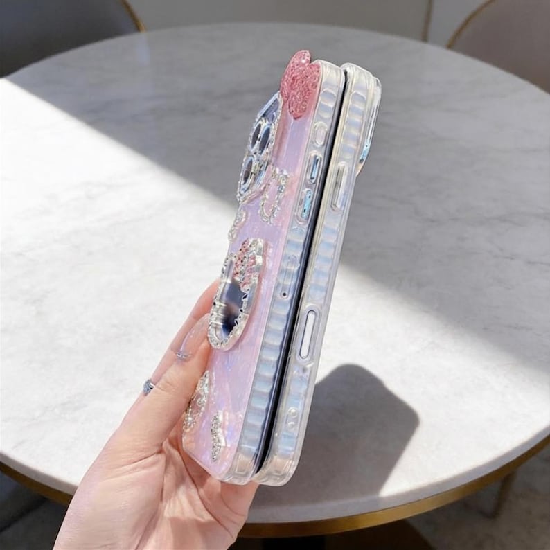 Pode incluir: Duas capas de telefone transparentes empilhadas. A capa interna &eacute; rosa claro com enfeites prateados, incluindo um la&ccedil;o, strass e formas decorativas. A capa externa &eacute; transparente.