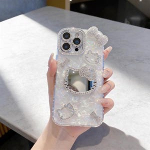 Pode incluir: Capa de telefone transparente com um acabamento brilhante e iridescente. A capa apresenta um design Hello Kitty com detalhes em strass, incluindo um grande rosto Hello Kitty com um espelho, la&ccedil;os e outras pequenas figuras Hello Kitty. A &aacute;rea da c&acirc;mera tamb&eacute;m &eacute; adornada com strass.