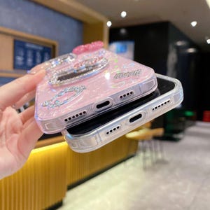 Pode incluir: Capa de telefone rosa com uma superf&iacute;cie brilhante e texturizada. A capa apresenta enfeites prateados, incluindo um suporte em forma de cora&ccedil;&atilde;o e detalhes decorativos. A capa &eacute; projetada para um smartphone e possui recortes para a c&acirc;mera e alto-falantes.