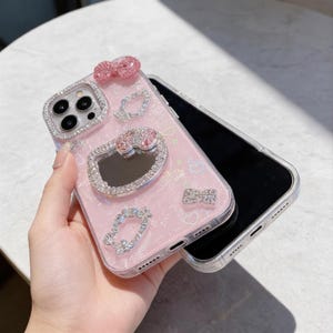 Pode incluir: Capa de telem&oacute;vel rosa com borda transparente, adornada com brilhantes strass. Apresenta um la&ccedil;o, um cora&ccedil;&atilde;o e outros elementos decorativos. A capa &eacute; projetada para um smartphone, oferecendo estilo e prote&ccedil;&atilde;o.