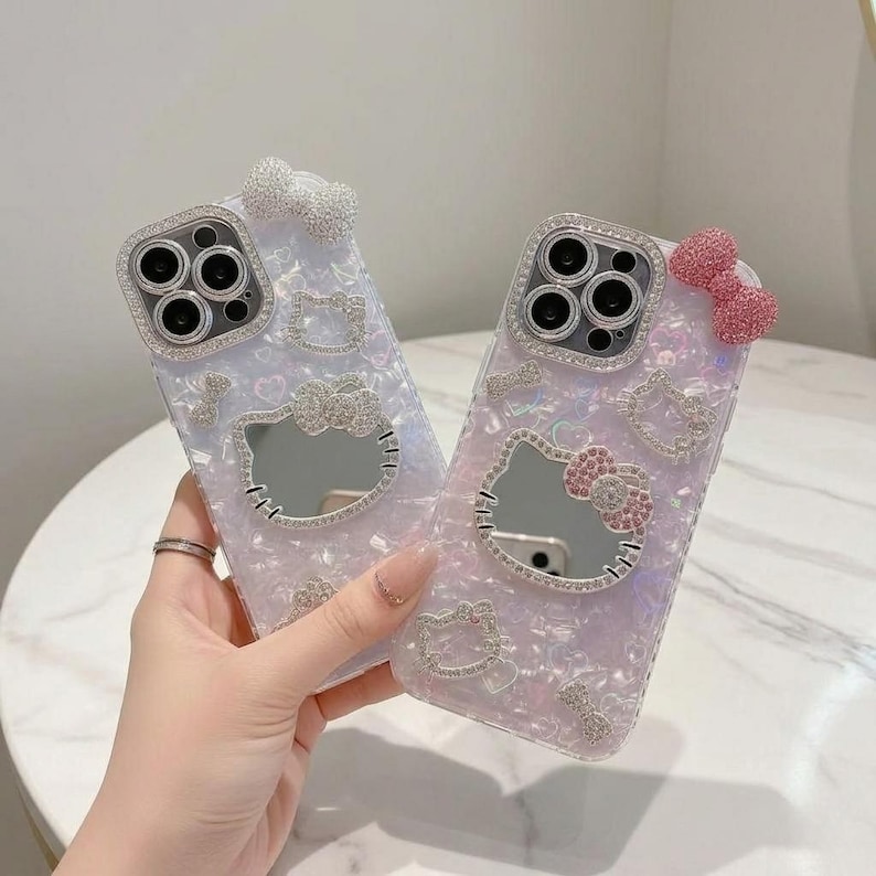 Pode incluir: Duas capas de telefone iridescentes com um design Hello Kitty. Cada capa apresenta um espelho em forma do rosto de Hello Kitty, adornado com strass. Uma capa tem um la&ccedil;o branco, a outra um la&ccedil;o rosa, ambos adornados com strass.