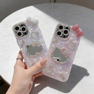 Pode incluir: Duas capas de telefone iridescentes com um design Hello Kitty. Cada capa tem um rosto Hello Kitty espelhado, adornado com strass e la&ccedil;os. A lente da c&acirc;mera &eacute; cercada por uma borda de strass. Um la&ccedil;o &eacute; branco, o outro &eacute; rosa.