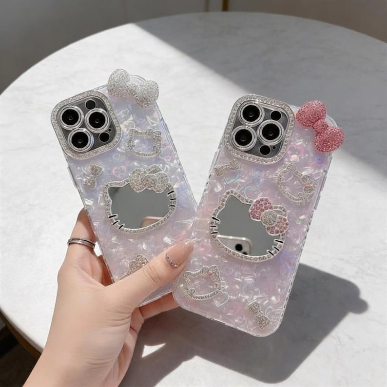 Pode incluir: Duas capas de telefone iridescentes com um design Hello Kitty. Cada capa tem um rosto Hello Kitty espelhado, adornado com strass e la&ccedil;os. A lente da c&acirc;mera &eacute; cercada por uma borda de strass. Um la&ccedil;o &eacute; branco, o outro &eacute; rosa.
