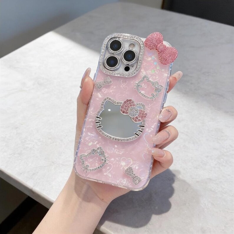 Pode incluir: Capa de telefone rosa com design Hello Kitty. A capa apresenta um espelho, um la&ccedil;o e strass brilhantes. A capa tem um acabamento perolado e &eacute; decorada com contornos de Hello Kitty.