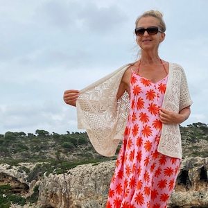 Puede incluir: Una mujer lleva un vestido maxi floral rosa y naranja con un cárdigan de ganchillo blanco. Está de pie en un acantilado rocoso con vistas a una masa de agua.
