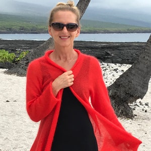 Könnte beinhalten: Eine Frau trägt einen roten Strickcardigan mit einem schwarzen Kleid darunter. Sie steht an einem Strand mit einer Palme im Hintergrund.