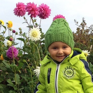 May include: A young person wearing a green knitted hat and a lime green jacket with black trim. The jacket has a white zipper and the words "Brooklyn 52" on the front. The person is standing in a garden with pink and white flowers.