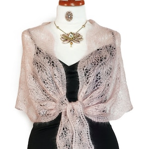 Puede incluir: Un chal de punto rosa delicado con un patrón de encaje. El chal está anudado en la parte delantera y cae sobre los hombros.