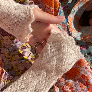 Pode incluir: Um close-up de uma peça de roupa de malha bege com um delicado padrão de renda. O tecido é macio e texturizado, com um brilho sutil. A peça está dobrada ao meio, revelando os detalhes intrincados do tricô.