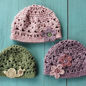 Könnte beinhalten: Drei gehäkelte Mützen in verschiedenen Farben mit Verzierungen. Die Mützen sind rosa, lila und grün. Die rosa Mütze hat eine Blumenverzierung, die lila Mütze hat zwei Blumenverzierungen und die grüne Mütze hat eine Schneckenverzierung.