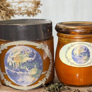 May include: Two amber glass jars filled with a golden substance, each featuring a circular label with an illustration of the Earth and the text Of Earth Apothecary and Health Market. The left jar has a black lid, while the right jar has a glass lid.