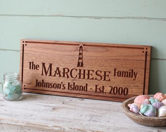 May include: A rectangular wooden sign with a dark brown finish featuring an engraved lighthouse icon and the text: The MARCHESE Family, Johnson's Island - Est. 2000. The sign is framed by a simple border with corner accents.