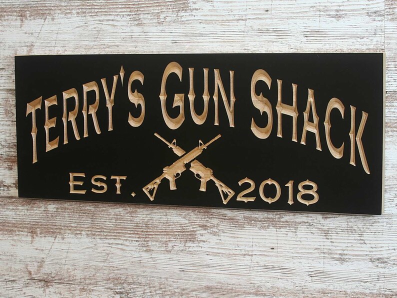 May include: A black wooden sign with the text "Terry's Gun Shack Est. 2018" in a light brown color. Two crossed rifles are depicted in the center of the sign.