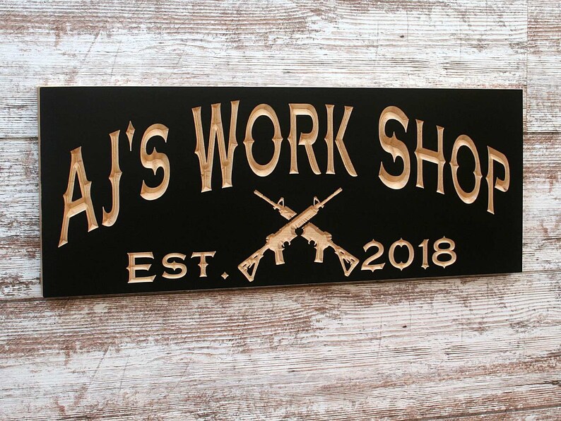 May include: Black wooden sign with the text "AJ's Work Shop Est. 2018" in a light brown color. Two crossed rifles are in the center of the sign.