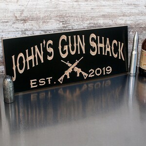 May include: Black wooden sign with the text "JOHN'S GUN SHACK EST. 2019" in a decorative font. Two crossed rifles are depicted in the center of the sign.