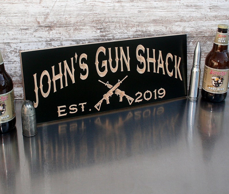 May include: Black wooden sign with the text "JOHN'S GUN SHACK EST. 2019" in a decorative font. Two crossed rifles are depicted in the center of the sign.