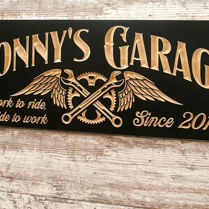 May include: Black wooden sign with gold lettering that reads "Jonny's Garage" with a winged wrench and gear design. The sign also says "Work to ride, Ride to work" and "Since 2018".