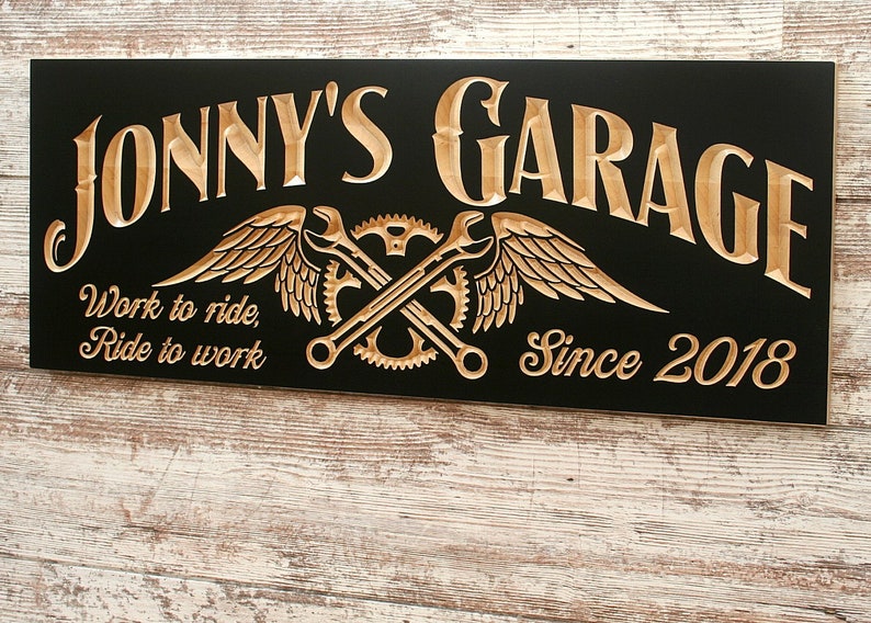 May include: Black wooden sign with gold lettering that reads "Jonny's Garage" with a winged wrench and gear design. The sign also says "Work to ride, Ride to work" and "Since 2018".
