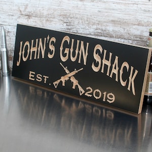 May include: Black wooden sign with the text "JOHN'S GUN SHACK EST. 2019" in a light brown font. Two crossed rifles are depicted in the center of the sign.