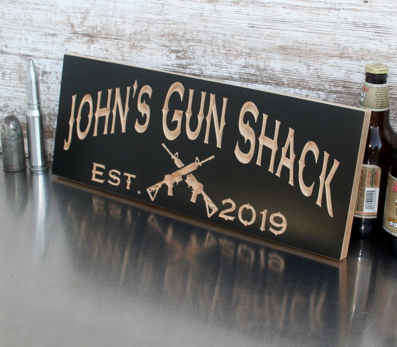 May include: Black wooden sign with the text "JOHN'S GUN SHACK EST. 2019" in a light brown font. Two crossed rifles are depicted in the center of the sign.