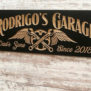 May include: Black wooden sign with gold lettering that reads "Rodrigo's Garage" with a pair of crossed wrenches and wings, and the text "Dad's Zone" and "Since 2018".