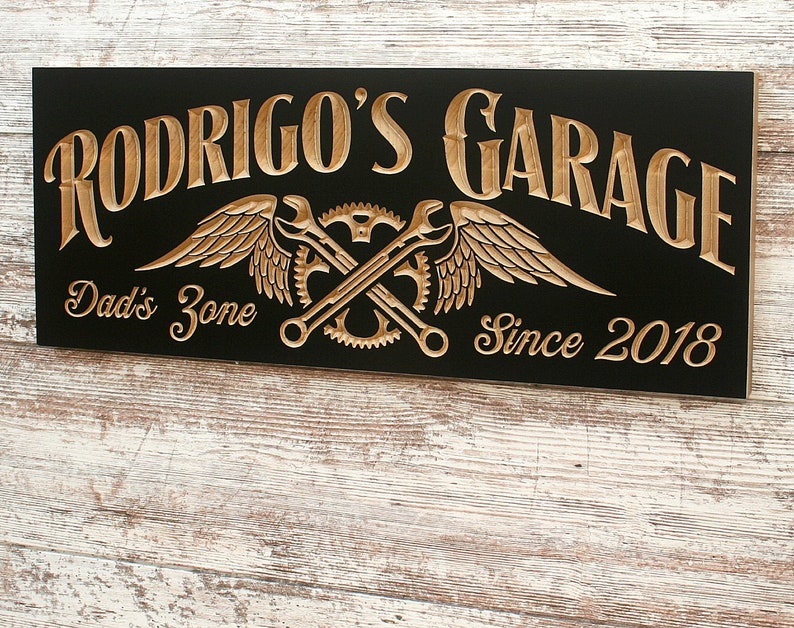 May include: Black wooden sign with gold lettering that reads "Rodrigo's Garage" with a pair of crossed wrenches and wings, and the text "Dad's Zone" and "Since 2018".