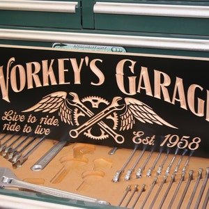 May include: A black wooden sign with the text "Workey's Garage" in brown lettering. The sign also features a winged wrench and gear design, the text "Live to ride, ride to live", and "Est. 1958".