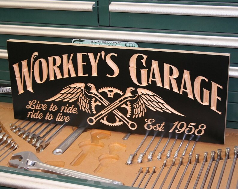 May include: A black wooden sign with the text "Workey's Garage" in brown lettering. The sign also features a winged wrench and gear design, the text "Live to ride, ride to live", and "Est. 1958".
