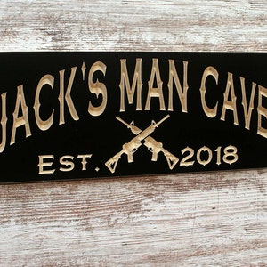 May include: Black wooden sign with gold lettering that reads "Jack's Man Cave Est. 2018" with two crossed rifles in the center.