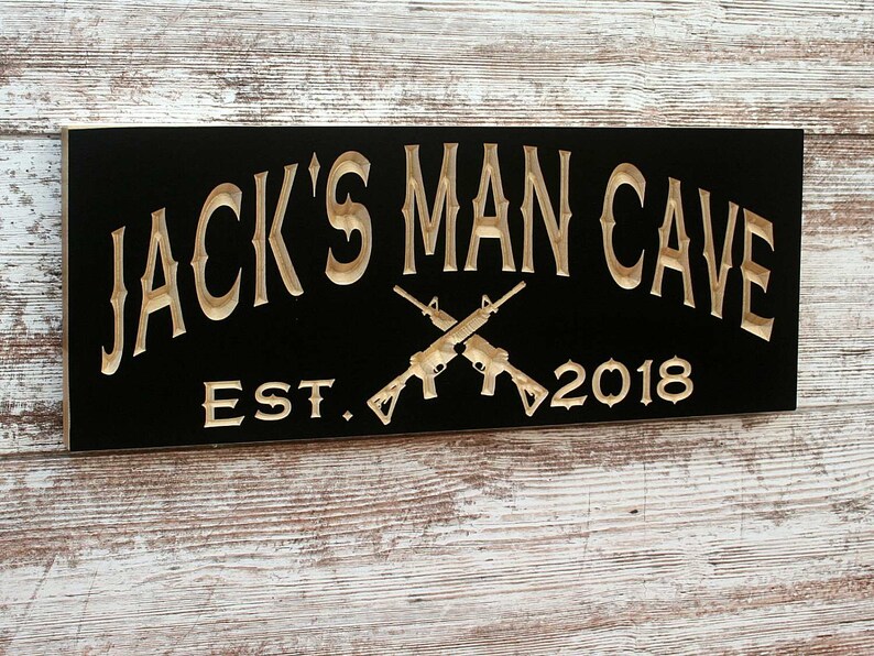 May include: Black wooden sign with gold lettering that reads "Jack's Man Cave Est. 2018" with two crossed rifles in the center.