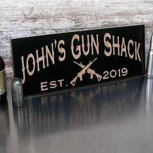 May include: Black wooden sign with the text "JOHN'S GUN SHACK EST. 2019" in a light brown font. Two crossed rifles are depicted in the center of the sign.