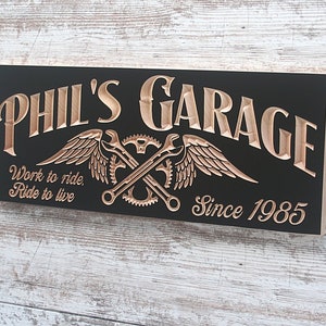May include: A black wooden sign with the words "Phil's Garage" in gold lettering. The sign also features a gold winged wrench and gear design and the text "Work to ride, Ride to live" and "Since 1985".