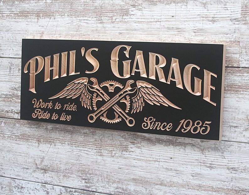 May include: A black wooden sign with the words "Phil's Garage" in gold lettering. The sign also features a gold winged wrench and gear design and the text "Work to ride, Ride to live" and "Since 1985".