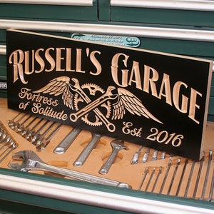 May include: A black wooden sign with the words "Russell's Garage" in a white, stylized font. The sign also features a brown, carved image of a pair of crossed wrenches with wings and gears, and the text "Fortress of Solitude Est. 2016".