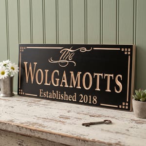 May include: A rectangular black wooden sign with carved light-colored lettering that reads The WOLGAMOTTS Established 2018. The sign features a decorative border with corner accents and is displayed horizontally on a distressed white surface.