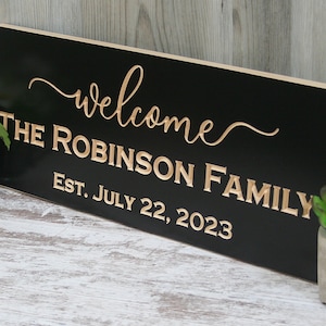 May include: Black wooden sign with gold lettering that reads "Welcome The Robinson Family Est. July 22, 2023".