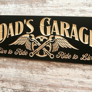 May include: Black wooden sign with the text "Dad's Garage" in gold lettering. The sign also features a gold winged design with crossed wrenches and gears. The text "Live to Ride" and "Ride to Live" is also in gold lettering.