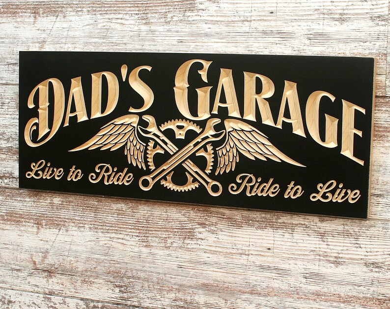 May include: Black wooden sign with the text "Dad's Garage" in gold lettering. The sign also features a gold winged design with crossed wrenches and gears. The text "Live to Ride" and "Ride to Live" is also in gold lettering.
