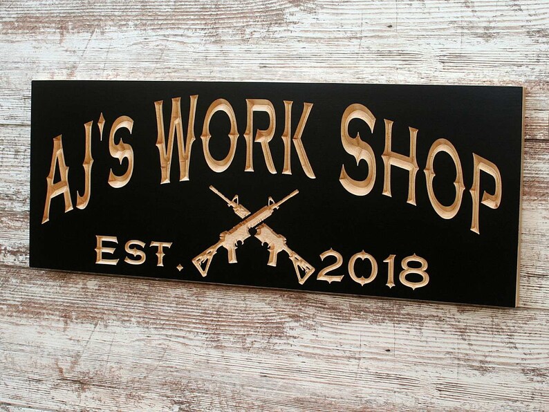 May include: A black wooden sign with the text "AJ's Work Shop Est. 2018" in gold lettering. Two crossed rifles are carved into the wood above the text.