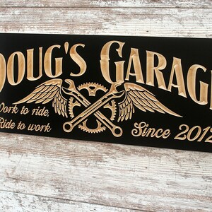 May include: A black wooden sign with the text "Doug's Garage" in a stylized font. The sign also features a pair of crossed wrenches with wings, gears, and the text "Work to ride, Ride to work" and "Since 2012".
