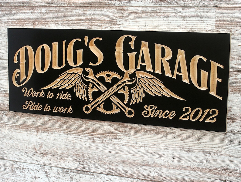 May include: A black wooden sign with the text "Doug's Garage" in a stylized font. The sign also features a pair of crossed wrenches with wings, gears, and the text "Work to ride, Ride to work" and "Since 2012".