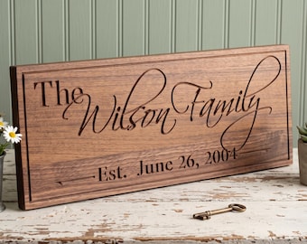 May include: A rectangular wooden sign with a dark brown finish featuring the engraved text The Wilson Family and Est. June 26, 2004. The sign has a carved border and is accompanied by a small metal key placed on the surface in front of the sign.