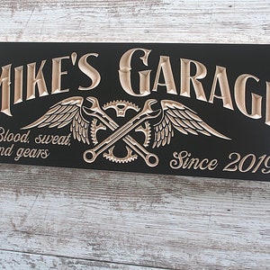 May include: Black wooden sign with the text "Mike's Garage" in a decorative font. The sign also features a pair of crossed wrenches with wings and gears, and the text "Blood, sweat, and gears" and "Since 2019".